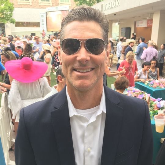 suited happy man wearing maverick sunglasses with people partying behind him