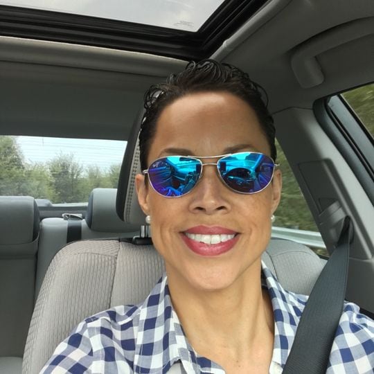 smiling woman with blue and white checkered shirt wearing blue lens sunglasses taking a selfie in car