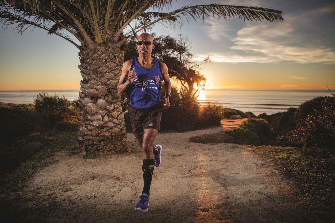 male runner running past palm tree wearing maui jim tank top and sunglasses with sunset in the background