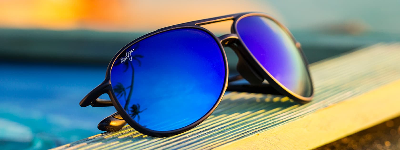 Sonnenbrille mit mattschwarzer Fassung und blauen Gläsern, in denen sich Himmel und Palmen spiegeln, auf Holz