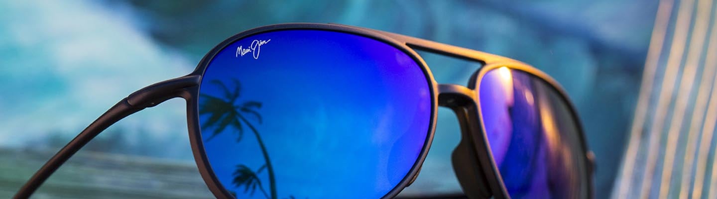 Sonnenbrille mit mattschwarzer Fassung und blauen Gläsern, in denen sich Himmel und Palmen spiegeln, auf Holz