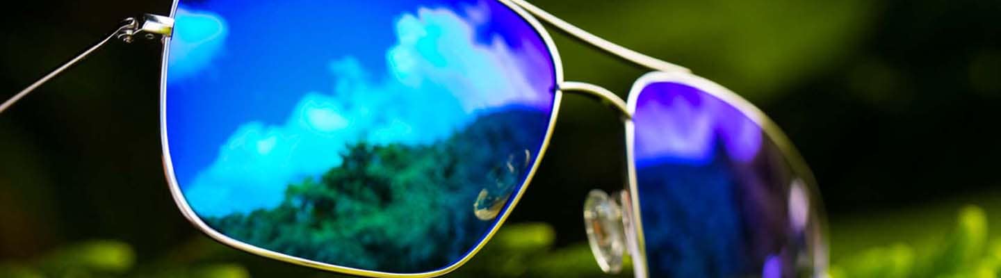 Sonnenbrille mit silberfarbener Fassung und blauen Gläsern, in denen sich Himmel und Palmen spiegeln