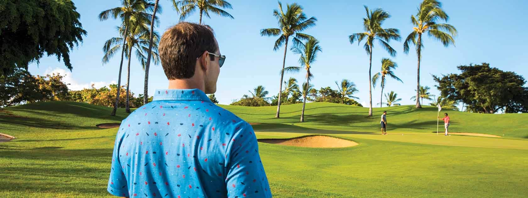 man with blue textured dress shirt standing on golf course with palm trees looking at other golfers