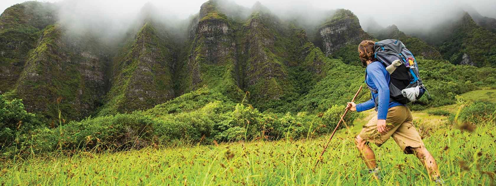 man backpacking through green field looking up at cloud covered rock face