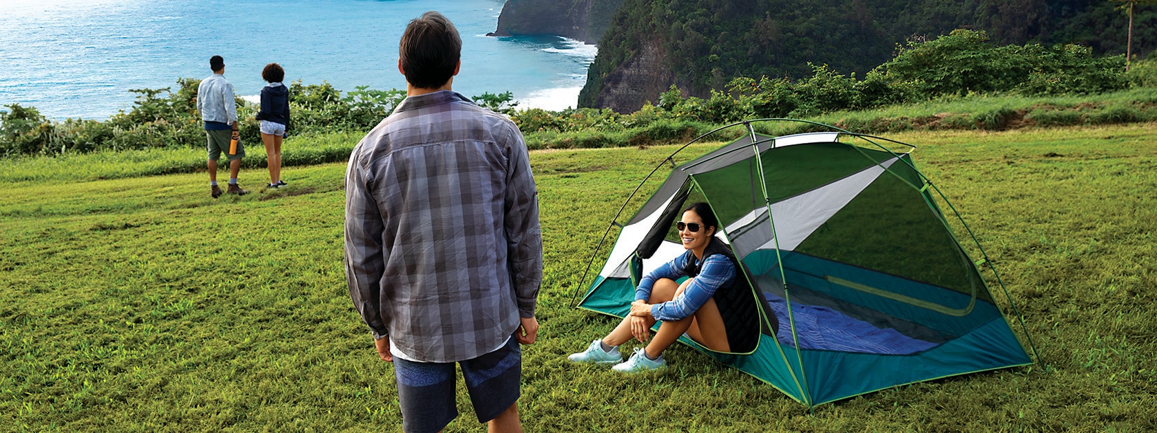 man standing next to woman sitting inside tent on a grassy hill overlooking ocean cliffside