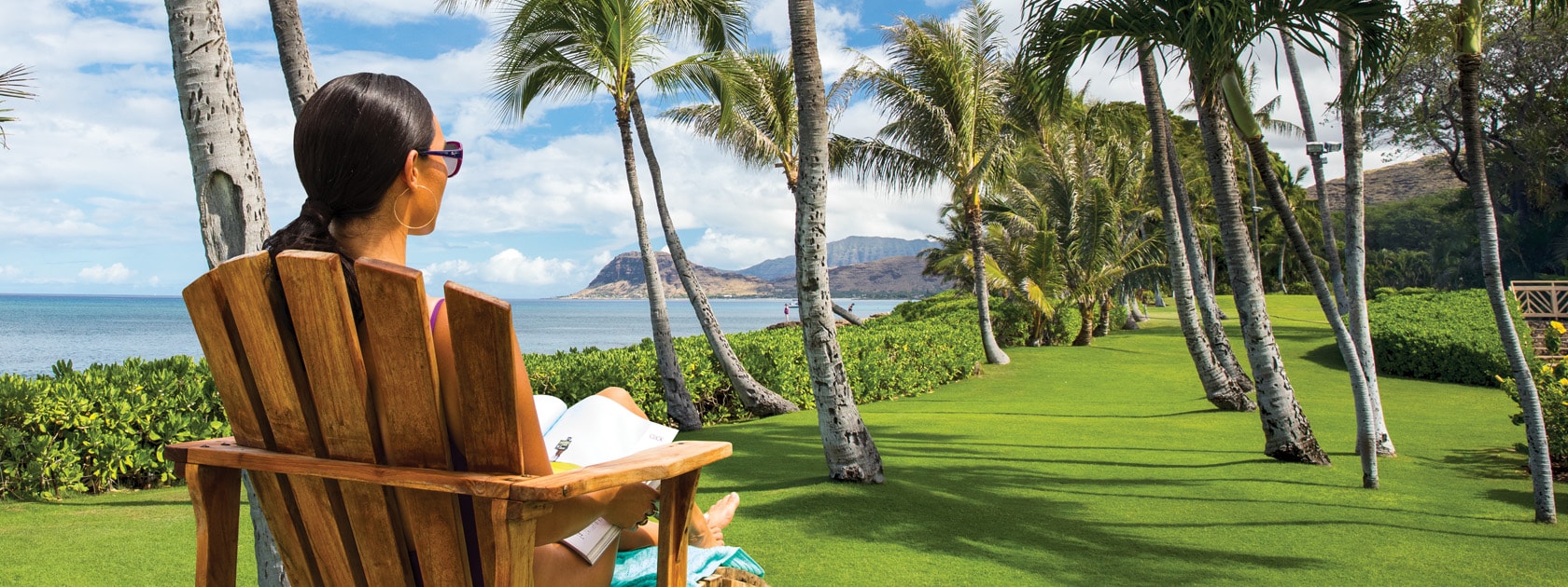 woman sitting in wood chair looking at sea side palm trees in green grassy field