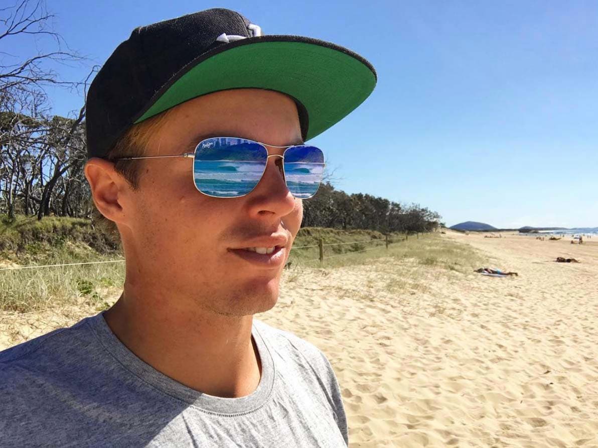 Mann mit Hut, grauem Hemd und Sonnenbrille, in deren Gläsern sich der Strand spiegelt