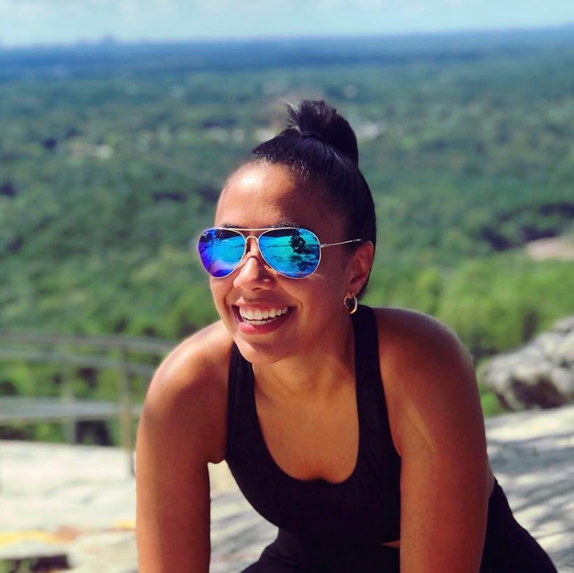 smiling woman with blue lens sunglasses sitting with hands on knees with forest behind her