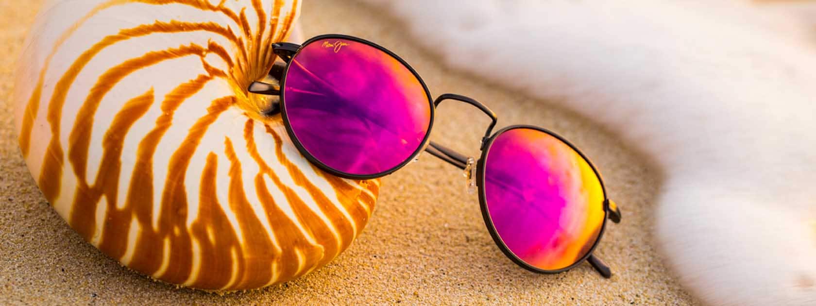 occhiali da sole con montatura metallica nera e lenti rosa appoggiati sulla sabbia e su conchiglie