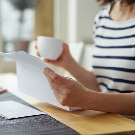 person holding cup of coffee reading paper at desk
