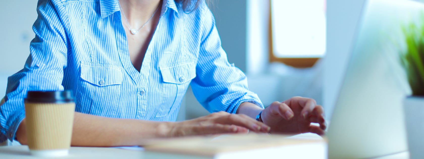 person with blue button up shirt sitting at table with cup of coffee typing on laptop computer