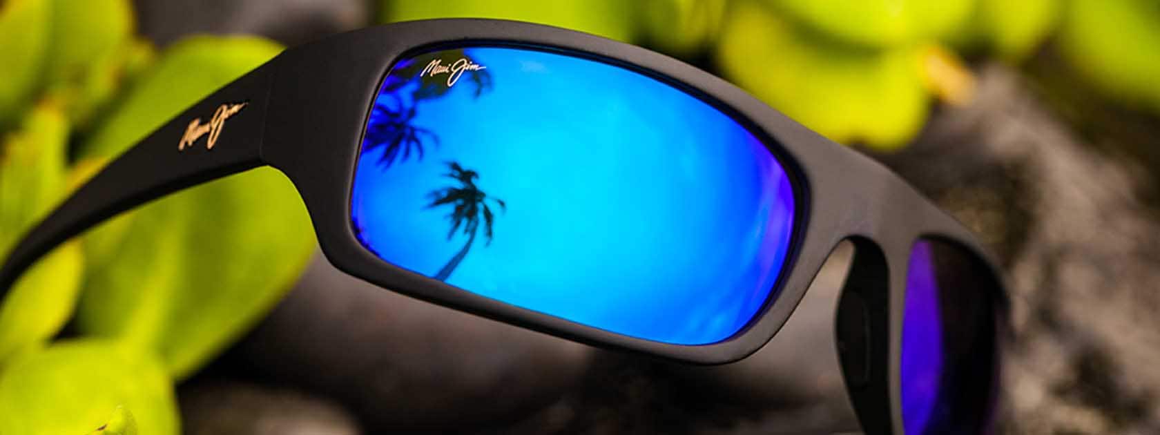 occhiali da sole con montatura nera opaca e lenti blu mostrati su foglie tropicali verdi