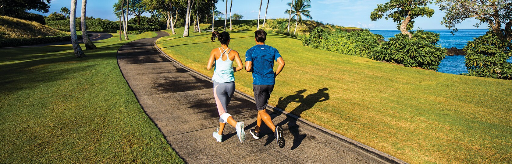 hombre y mujer corriendo por un camino junto a un césped verde con el océano y palmeras al fondo
