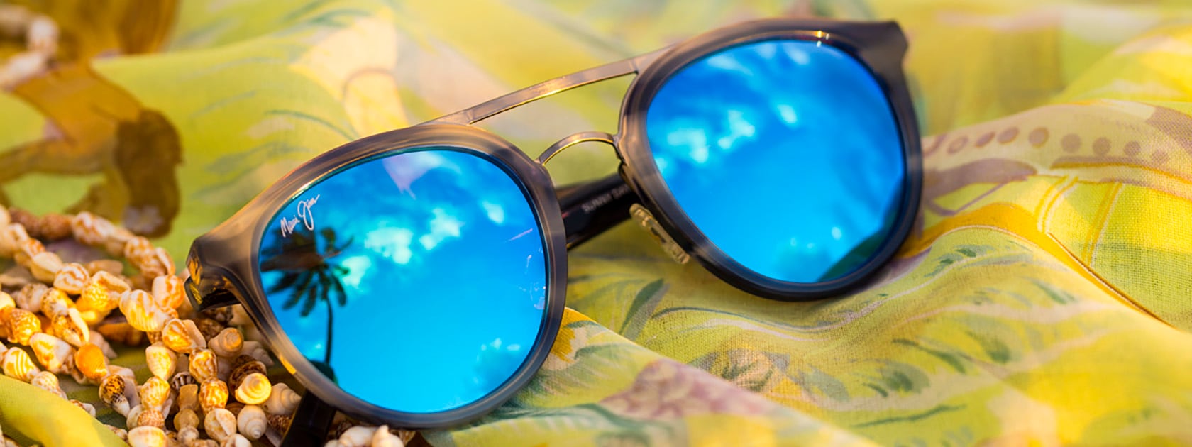 Braun gestreifte Sonnenbrillenfassung mit blauen Gläsern auf gelbem, blumigen Hintergrund und Muschelkette